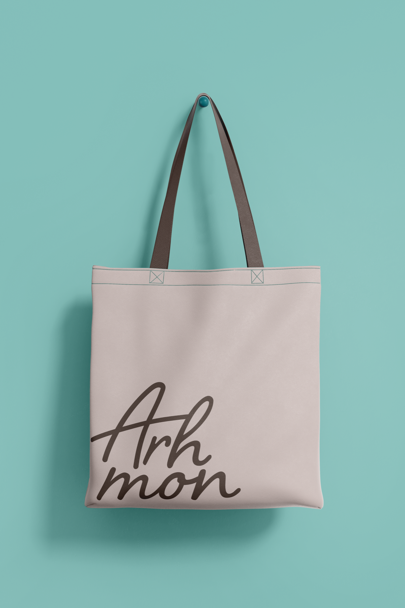 Intended application mockup: a blush tote bag with brown straps hanging on a teal wall, printed with the Arhmon handwritten lettering.