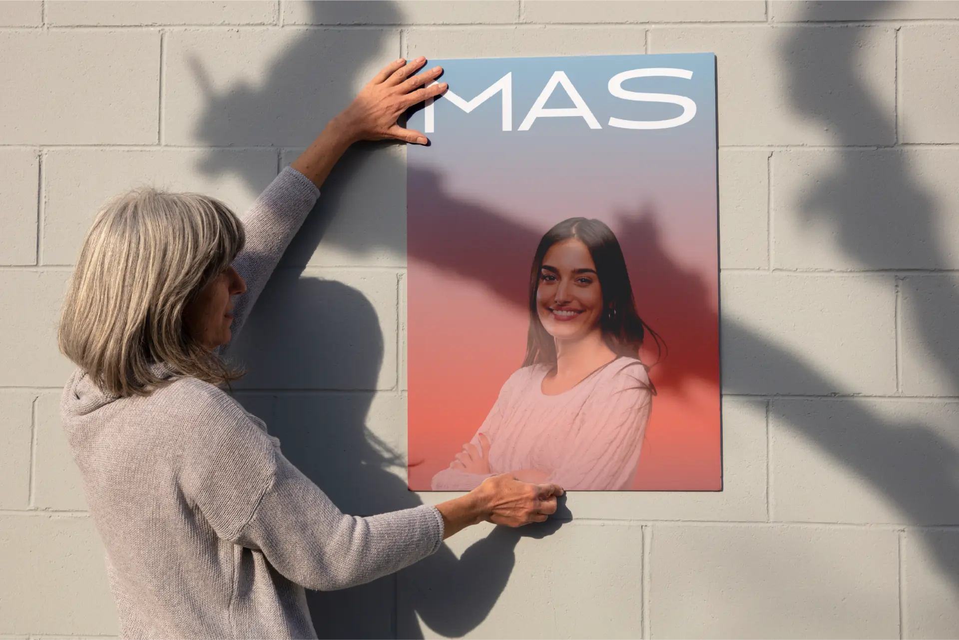 Brand application mockup: a woman mounts a large MAS poster on a painted cinder block wall; the poster shows the white MAS wordmark above a portrait on a blue-to-coral gradient.