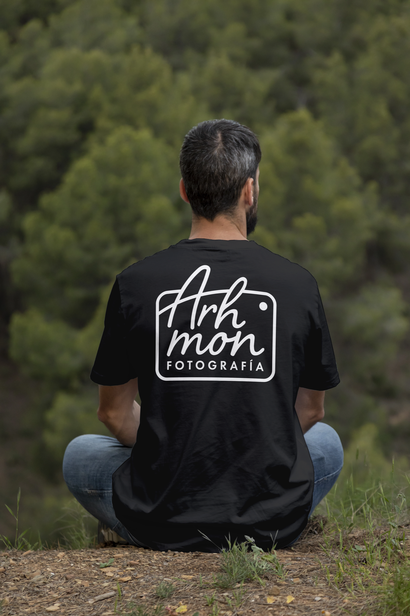 Intended application mockup: a seated person seen from behind wearing a black t-shirt with the large white Arhmon Fotografía camera-frame mark across the back.