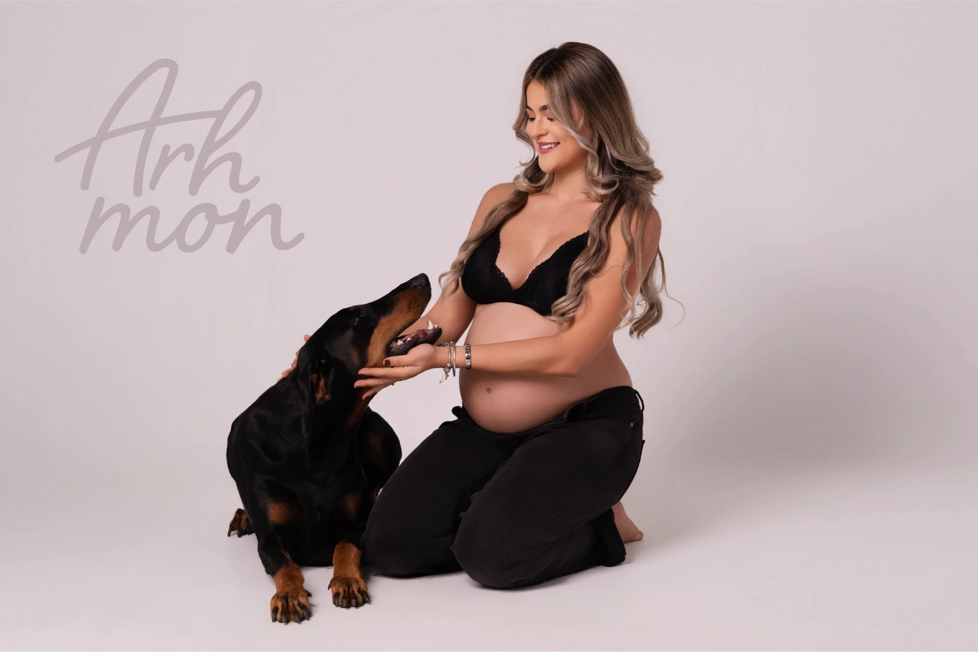 A maternity portrait of a pregnant person kneeling beside a Doberman dog on a soft neutral backdrop, with the handwritten Arhmon mark in the upper-left corner.